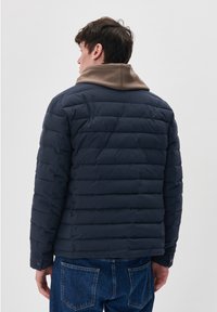 Mornarska pufastojna jakna z zaključenim dizajnom, ki ima kontrastno rjavo kapuco. Material se zdi gladek, s pritrdilnimi gumbi na manšetah.