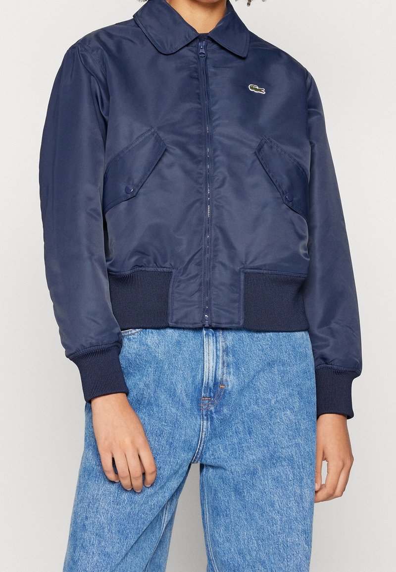 Personne portant une veste bleu marine zippée avec des poches à rabats boutonnées et des poignets côtelés, associée à un jean en denim bleu clair.