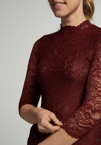Bordeaux kanten top met lange mouwen, hoge hals en bloemenpatronen. Heeft een geschulpte rand aan de mouwen, wat een delicate textuur laat zien.