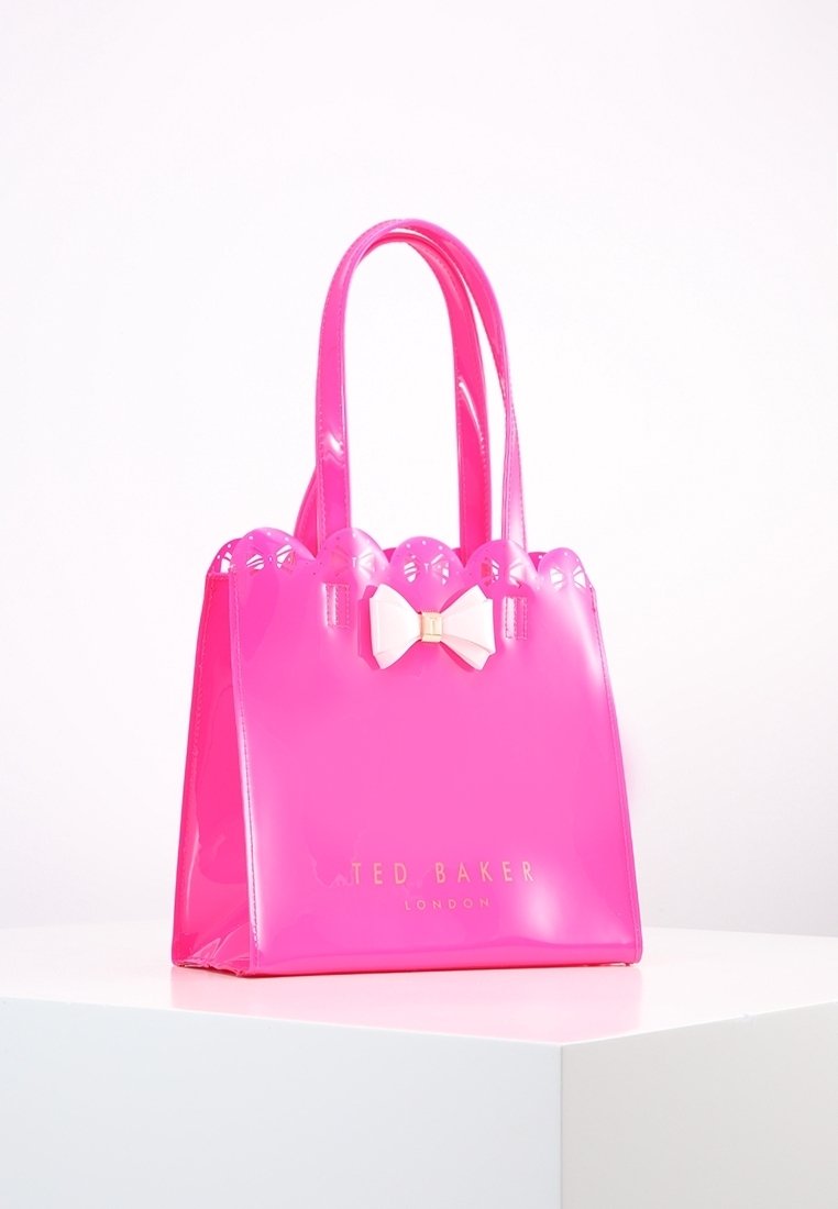 Sac à main Ted Baker rose vif avec bord supérieur festonné, double poignée et nœud blanc décoratif à l'avant, présenté sur une surface blanche.