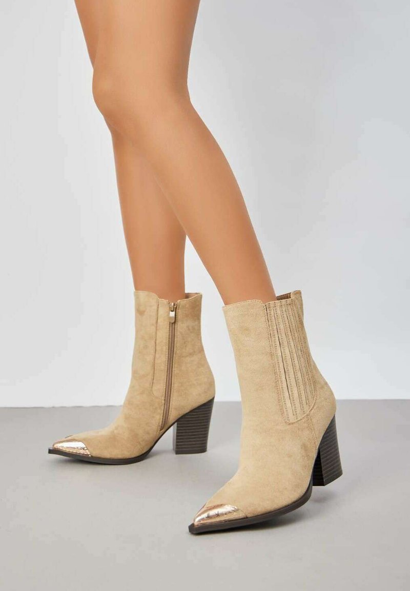 Bottes chelsea en suède beige avec un bout pointu et des accents métalliques. Équipées d'une fermeture éclair sur le côté et d'un design côtelé sur le haut de la tige. Talon épais.