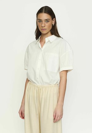 Hosszú barna hajú nő ezüst karika fülbevalóval, laza, fehér rövid ujjú, gombos inggel és bézs, húzózsinóros nadrággal, fehér háttér előtt áll.