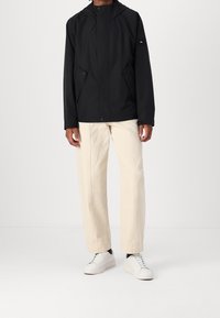 Czarna kurtka z kapturem o gładkiej fakturze, zapinana na zatrzaski, z bocznymi kieszeniami, zestawiona z jasnokrémowymi spodniami i białymi trampkami.