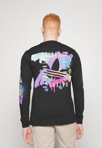 Mężczyzna z krótkimi kręconymi rudymi włosami, ubrany w czarną koszulę z długim rękawem z kolorowym, abstrakcyjnym wzorem Adidas na plecach oraz beżowe spodnie.