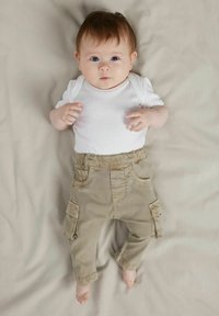 Valge lühikeste varrukatega bodi, mis on kombineeritud beežide taskutega karmiinpükstega. Väike laps on beežil taustal, hele nahk ja pruunid juuksed.