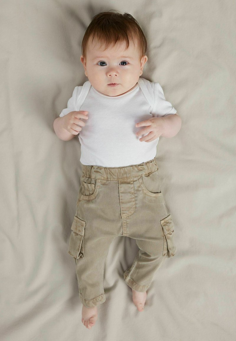 Valge lühikeste varrukatega bodi, mis on kombineeritud beežide taskutega karmiinpükstega. Väike laps on beežil taustal, hele nahk ja pruunid juuksed.