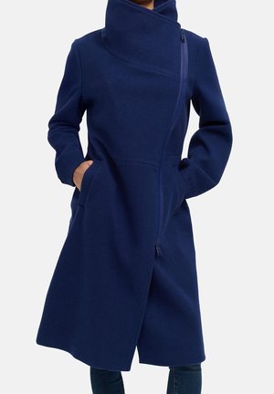 Woman wears knee-length navy blue coat with high collar, asymmetrical front zipper, and hands in side pockets.