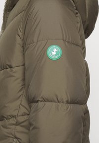 Rękaw oliwkowo-zielonej puchowej kurtki z okrągłą zieloną naszywką przedstawiającą białą kaczkę oraz napis "Save The Duck".