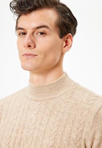 Beige stickad tröja med hög krage, med en texturerad kabeldesign. Materialet verkar mjukt och lätt.