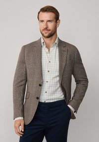 Blazer a quadri marrone con revers, due bottoni e tasche frontali, abbinato a una camicia a righe bianche e a un pantalone blu navy.
