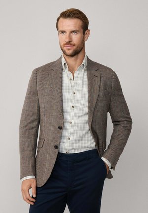 Blazer a quadri marrone con revers, due bottoni e tasche frontali, abbinato a una camicia a righe bianche e a un pantalone blu navy.