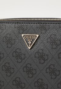 Portefeuille noir texturé arborant un logo triangulaire doré avec "GUESS USA" et un point d'interrogation, rehaussé d'une fermeture éclair dorée.