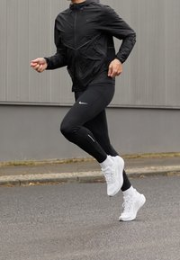 Czarna kurtka przeciwdeszczowa Nike, dopasowane czarne legginsy i białe buty sportowe. Teksturowany materiał i odblaskowe akcenty w odzieży sportowej.