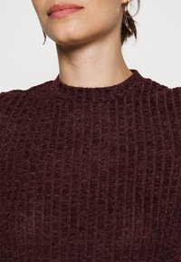 Femme portant un pull en maille côtelée bordeaux avec un col rond monté et de longues boucles d'oreilles torsadées dorées en forme d'anneau.