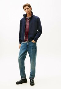 Jacket a zip con cappuccio blu navy sopra una maglietta a righe bordeaux e bianche, abbinata a jeans in denim azzurri e scarpe nere. Design semplice, vestibilità casual.