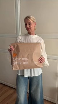 Bruine papieren tas met witte tekst "Hou van mij. Draag mij." en een oranje afbeelding, vastgehouden door een persoon in een witte blouse en uitzwenkende blauwe jeans.