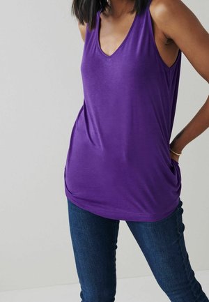 Mujer con una camiseta sin mangas de cuello en V morada y jeans azul oscuro, posando con una mano en la cadera frente a un fondo liso.
