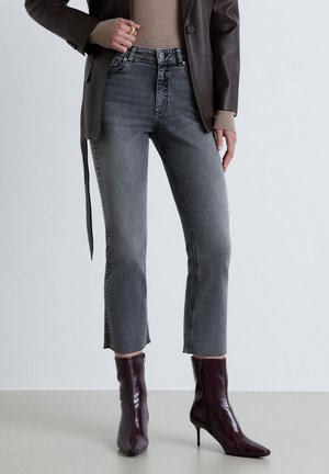 Femme portant un jean gris foncé court avec un ourlet brut, des bottines en cuir marron à talons, un pull beige et une veste en cuir marron foncé.