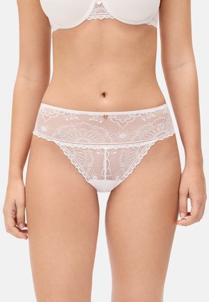 Mujer con conjunto de lencería blanca de encaje, mostrando diseño floral de encaje en las bragas de cintura alta y el sujetador a juego sobre un fondo claro.