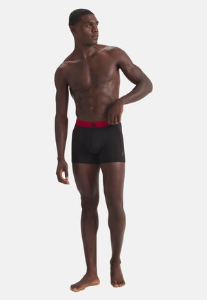 Athletischer Mann steht barfuß, trägt schwarze Boxershorts mit rotem Bund und schaut nach links vor einem schlichten Hintergrund.