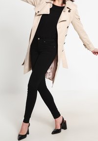 Manteau beige avec des boutons noirs et des poignets ajustables, porté par-dessus un haut noir et un jean skinny noir. Des chaussures à talons noires complètent la tenue.