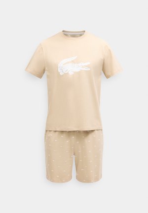 Bež kratka majica s kratkimi rokavi z velikim belim logotipom krokodila in ujemajočimi kratkimi hlačami z majhnimi belimi vzorci krokodila.