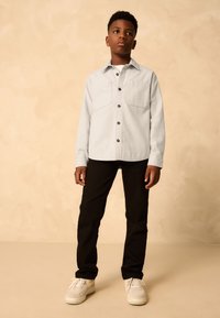 Light gray shirt with two front pockets, black buttons, and long sleeves. Paired with black pants and white sneakers. Neutral background.