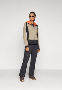 Wandeljack in beige en marineblauw met een oranje fleece-gevoerde capuchon, gecombineerd met zwarte broek en bruine wandelschoenen. Zonnebril maakt de look compleet.