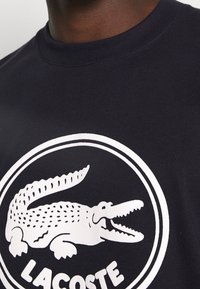Granatowa bawełniana koszulka z dużym białym logo krokodyla oraz napisem "LACOSTE" w okrągłym obrysie na wysokości klatki piersiowej.