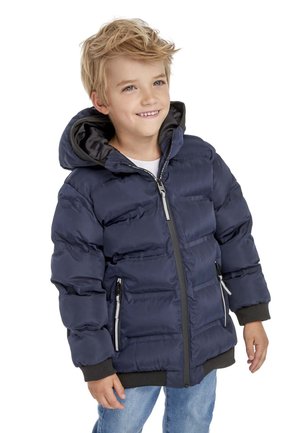 Un jeune garçon souriant avec des cheveux blonds en désordre portant une veste matelassée bleue marine avec capuche et un jean bleu clair sur fond blanc.