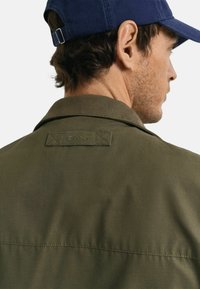 Olive green jacket with a collar and GANT logo patch on the back. Features smooth texture and consistent stitching detail.
