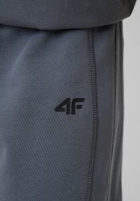 Gros plan sur un pantalon en tissu gris avec logo noir "4F" et couture visible sur le côté.