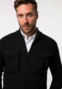 Schwarze Zip-Up-Strickjacke mit gerippter Textur, ausgestattet mit zwei Brusttaschen. Darüber wird ein weißes Hemd mit Kragen getragen. Einfaches, funktionales Design.