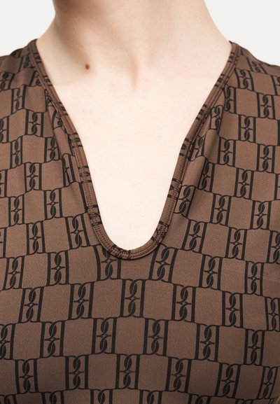 Άτομο που φοράει καφέ μπλούζα με V λαιμόκοψη και επαναλαμβανόμενο μαύρο γεωμετρικό μοτίβο, με ορατούς τον λαιμό και το άνω μέρος του στήθους.