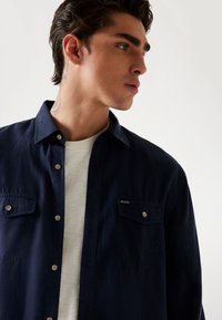 Marineblaues Hemd mit Knopfleiste, zwei Brusttaschen und Logodetail. Getragen über einem weißen Rundhalsshirt. Glattes Material und taillierte Passform.