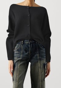 Musta, nappikiinnitteinen neule, jossa olkapäät paljastava muotoilu, pitkät hihat ja resorikuvio, yhdistetty haalistuneisiin sinisiin farkkuihin.