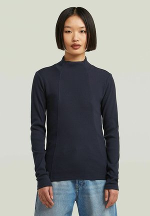 Femme avec des cheveux noirs raides portant un haut ajusté à manches longues bleu marine et un jean bleu clair sur un fond clair.