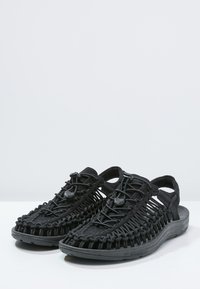 Black sandals with woven textile upper, adjustable lacing system, and open back design. Features a flexible rubber sole and smooth finish.