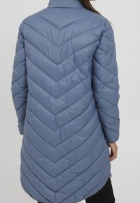 Vadderad lång kappa i mjuk blå färg med zigzag-quiltad design, hög krage och långa ärmar. Har sidofickor och en något rundad nederkant.