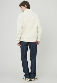 Pull en crème, texturé, avec un col montant, associé à un jean bleu foncé. Le mannequin fait face à l'arrière, mettant en valeur le matériau en maille douce.