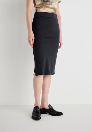 Jupe midi noire et ajustée avec une petite fente latérale, portée avec un haut beige à motif marron et des chaussures plates slip-on noires à talons bas.