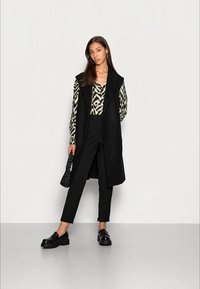 Colete longo preto sobre uma blusa preta e branca com padrão, calças pretas e sapatos pretos robustos. Mala texturizada, acessórios mínimos.