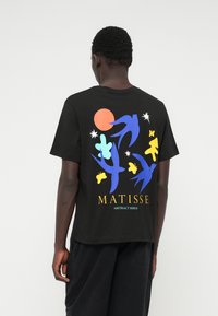 Fekete pamut póló, hátán absztrakt grafikával, kék, sárga és zöld madarakkal és formákkal, feliratozva: "MATISSE ABSZTRAKT MADARAK."