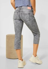 Person wearing gray cropped jeans and beige sneakers standing near a beige cylindrical textured stool on a light floor and wall.