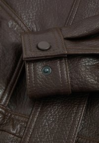 Pruun nahast rihm, millel on tekstureeritud pind, musta nööbiga ja täpsete õmbluse detailidega.