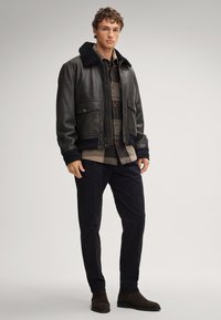 Czarna skórzana kurtka bomberka z kołnierzem z baranka, pod spodem zapinana na guziki koszula w kratę oraz ciemne spodnie. Stylizację dopełniają brązowe zamszowe botki za kostkę.