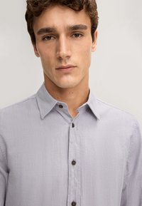Camicia button-up grigia chiara con un finish testurizzato, dotata di colletto a punta e bottoni marroni, sapientemente tagliata per un aspetto ordinato.