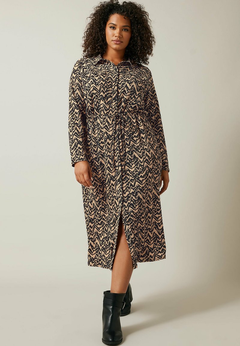 Langes, gemustertes Kleid mit einem schwarzen und beige Zickzackdesign, kragenlosem Ausschnitt und Kordelzugtaille. Gespalterter Saum und kombiniert mit schwarzen Stiefeln.