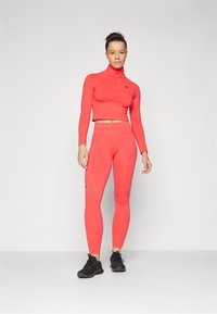To-delt treningsantrekk i livlig korall, bestående av en tettsittende helårsjakke med glidelås og matchende høytaljede tights. Strukturert stoff.