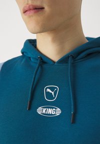 Felpa teal in tessuto morbido con cappuccio e cordino, patch del logo Puma e logo testuale 'KING', con accenti a contrasto sulle maniche.